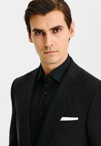 Czarna marynarka o satynowym wykończeniu, z białą poszetką i czarną koszulą pod spodem. Krój jest dopasowany i zorganizowany.