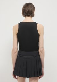 Persona con capelli corti e marroni che indossa un top senza maniche nero e una gonna plissettata nera, vista di schiena su uno sfondo semplice e chiaro.