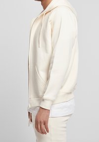 Sudadera con cremallera de color crema claro, con capucha ajustable, bolsillos frontales y puños acanalados. Textura suave y lisa; corte relajado.
