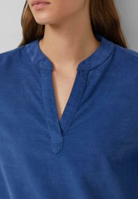 Haut en tissu bleu avec un col mandarin, présentant un décolleté en encolure angle et une texture subtile. Finition lisse, design décontracté.