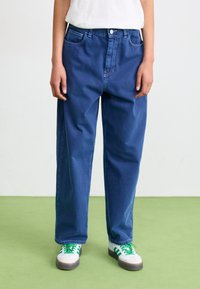 Blauwe denim jeans met een ontspannen pasvorm, voorzien van een knoopsluiting, vijf zakken en een licht vervaagde afwerking. Gecombineerd met sneakers met groene accenten.