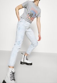 Persona vistiendo jeans azul claro, botas de plataforma blancas con cordones y una camiseta gris con el gráfico "RUSH", posando frente a un fondo blanco liso.