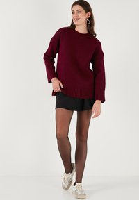 Burgundi színű, oversized kötött pulóver bordás mandzsettákkal és aljjal, fekete szoknyával és áttetsző fekete harisnyával viselve, világos sportcipőkkel párosítva.