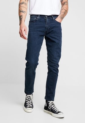 Jeans bleu foncé coupe slim, avec cinq poches, passants de ceinture et une texture denim lisse ; portés avec des baskets montantes noires.