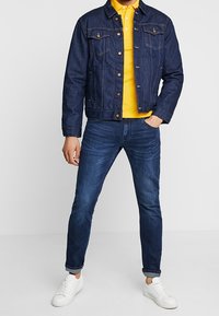 Kurtka dżinsowa w ciemnoniebieskim kolorze z przednimi kieszeniami, żółta koszulka polo, ciemne dżinsy lekko zwężane oraz białe trampki. Prosty, casualowy wygląd.