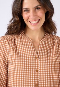 Blusa a scacchi marrone e bianca con scollo rotondo, chiusura con bottoni e maniche arricciate, realizzata in tessuto leggero.