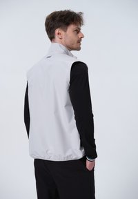 Lys grå ærmeløs vest med høj krave, glat tekstur, elastisk kant og logo på ryggen, båret over en sort langærmet skjorte.