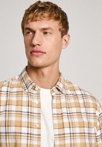 Camicia a maniche corte a quadri beige e bianca con dettagli blu, realizzata in tessuto morbido. Presenta un colletto button-down e una tasca applicata.