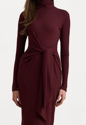 Vrouw draagt een lang bordeauxrode jurk met lange mouwen en een coltrui, met een grote stoffen knoop vastgebonden rond de taille.