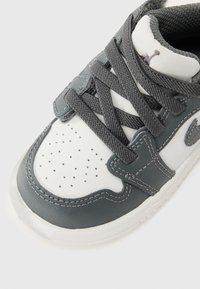 Baskets grises et blanches avec un dessus en cuir texturé, une pointe perforée et des lacets gris. Présente un logo et des accents de couture renforcée.