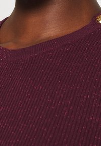 Bordeauxrode geribbelde trui met een getextureerd oppervlak en een subtiele glans, voorzien van een ronde halslijn en een decoratieve gouden knoop op de schouder.