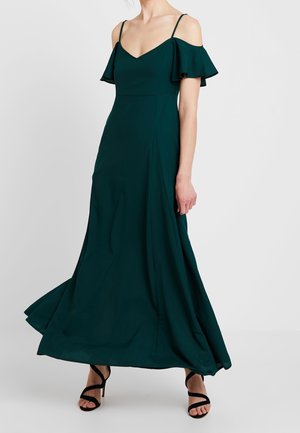 Kobieta w długiej sukience maxi w ciemnozielonym kolorze, z odkrytymi ramionami i cienkimi ramiączkami, nosząca czarne sandały na wysokim obcasie na jednolitym tle.