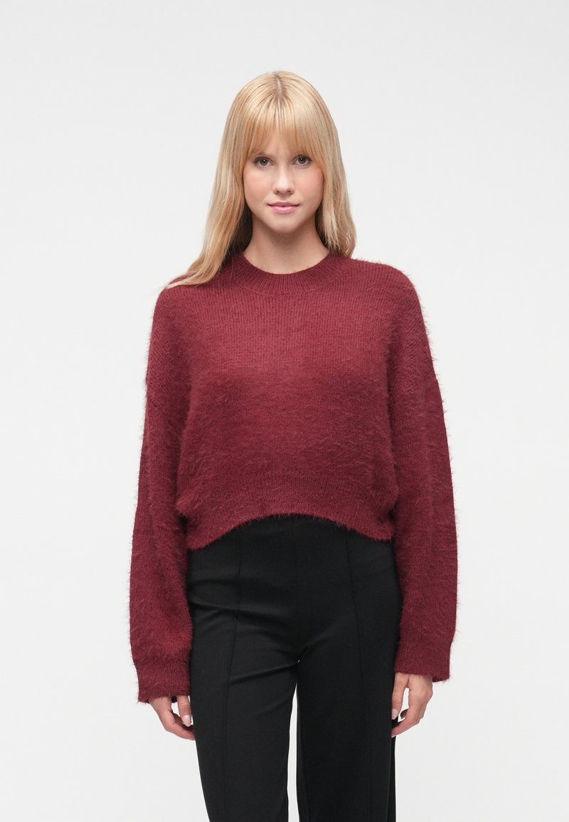 Μπορντό cropped πουλόβερ με αδρή υφή και μακριά μανίκια, με στρογγυλή λαιμόκοψη και ριμπ μανσέτες. Συνδυασμένο με μαύρο παντελόνι.