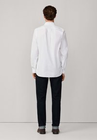 Camisa blanca de manga larga con un pliegue en la espalda, combinada con jeans de mezclilla oscuros arremangados en los puños y zapatos marrones, vista desde la parte trasera.