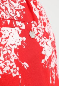 Rode stof met witte bloemenprint en een klein rond zilveren metalen label dat nabij een naad is bevestigd.