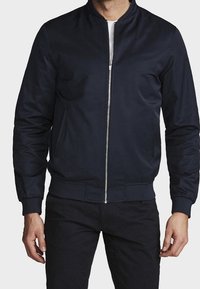 Chaqueta bomber azul marino hecha de tela lisa, con cremallera frontal, cuello y puños acanalados. Se pueden ver dos bolsillos laterales.