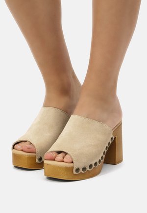 Zuecos abiertos de ante beige con un tacón de madera grueso. Presentan un borde festoneado y tachuelas circulares a lo largo del lado.