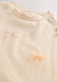 Tela color crema con dos pequeños lazos de tela transparente, uno rosa pálido y otro melocotón claro, cerca del escote redondo.