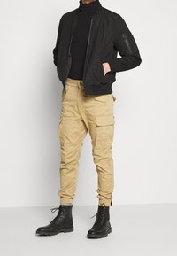 Svart bomberjacka över en svart polotröja, kombinerad med beige cargobyxor och svarta snörstövlar. Texturerat material med flera fickor.