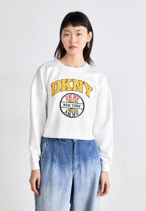 Hvit cropped sweatshirt med dristig gul "DKNY" logo og rund grafikk. Ribbestrikkede mansjetter og myk stoff; parret med lyseblå denim bukser.