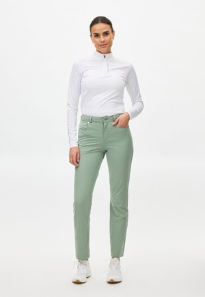 Camisa blanca de manga larga con cremallera combinada con pantalones ajustados verde claro. La modelo lleva zapatillas blancas. El conjunto tiene una textura suave y un diseño minimalista.