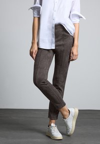 Suède-achtige bruine broek met zijdelingse naaddetails, gecombineerd met een wit overhemd met knopen en lichtgrijze sneakers. Soepele textuur; getailleerd ontwerp.
