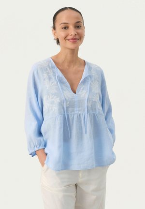 Blusa in lino azzurro chiaro con scollatura a V, maniche a tre quarti e ricami floreali bianchi sul colletto. Vestibilità comoda con vita arricciata.