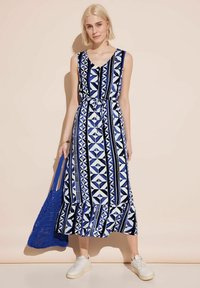 Robe maxi sans manches en bleu, blanc et noir avec des motifs géométriques ; elle dispose d'une taille à cordon de serrage et d'un ourlet festonné. Associée à un sac en crochet bleu.