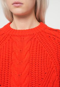 Punane kudumiga sviiter ümmarguse kaelusega ja palmiku mustriga. Omab soonikvarrukaid ja tekstureeritud pinda, esitledes elavat värvi.