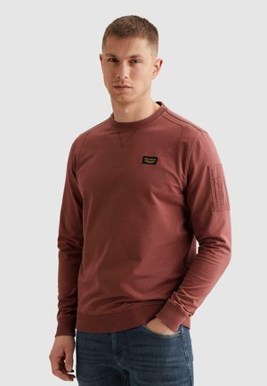 Mężczyzna z krótkimi włosami, ubrany w rdzawobrązową bluzę z długim rękawem z małą naszywką na piersi oraz niebieskie dżinsy, stojący na tle jasnoszarego tła.