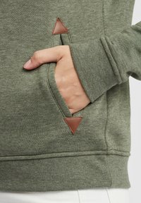 Green sweatshirt with a textured finish, featuring a visible hand in a pocket with a triangular brown leather accent.