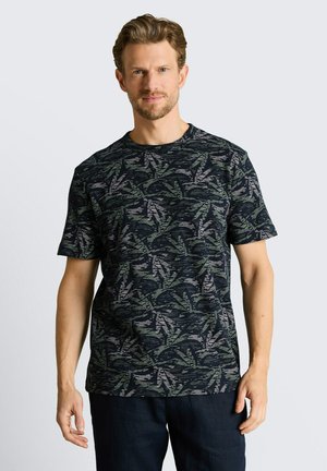 Man met kort krullend haar, die een donker T-shirt met korte mouwen en grijze en groene bladpatronen draagt, staand tegen een lichte achtergrond.