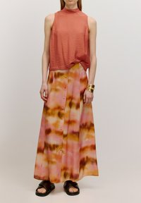 Roodbruine mouwloze top met een hoge hals, gecombineerd met een lange tie-dye rok in roze, oranje en bruin, met knoopdetails.