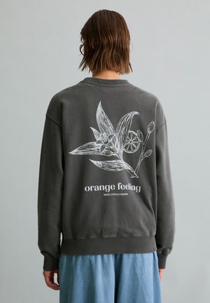 Persoon met kort bruin haar draagt een donkergrijze trui met witte botanische print en tekst "orange feeling" op de rug, gecombineerd met lichtblauwe broek.