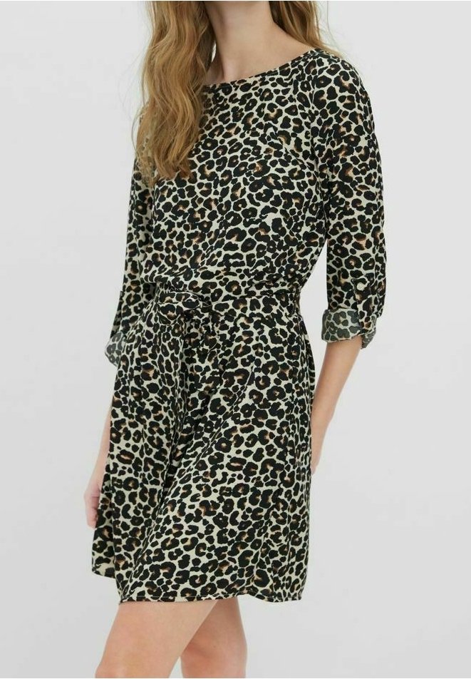 Femme portant une robe mi-longue à manches longues imprimée léopard avec une ceinture à la taille, debout avec un bras légèrement derrière le dos.