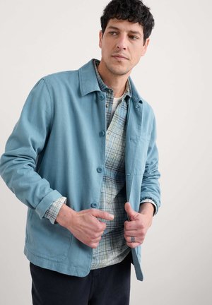 Light blue button-up jacket in soft fabric features a collared neck, front pockets, and a relaxed fit, layered over a checkered shirt.