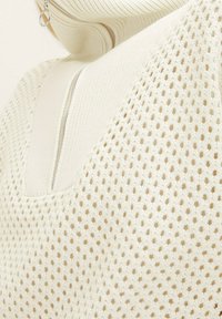 Maglione color crema chiaro con un lavorazione testurizzata simile a una rete. Presenta una lampo parziale e un colletto alto e ripiegato. Motivo discreto su tutta la superficie.