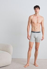 Šedé bavlnené boxerky s čiernym pásom s logom Nike, priliehavý dizajn a hladká textúra. Model stojí naboso na jednoduchom pozadí.