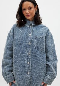 Blaue Jeanshemd mit einer strukturierten Webart, großen Knöpfen, einem Kragen und einer Brusttasche, hat einen oversized Schnitt und lange Ärmel.