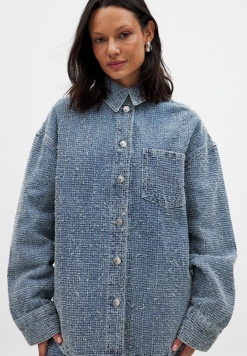 Blaue Jeanshemd mit einer strukturierten Webart, großen Knöpfen, einem Kragen und einer Brusttasche, hat einen oversized Schnitt und lange Ärmel.