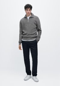 Maglione a coste grigio con collo con zip, indossato sopra una t-shirt bianca, abbinato a pantaloni scuri e sneakers bianche. Design semplice e moderno.