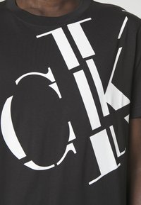 Svart T-shirt i bomull med framträdande vit grafisk text. Texten har varierande storlekar och stilar, vilket skapar en djärv och modern design.