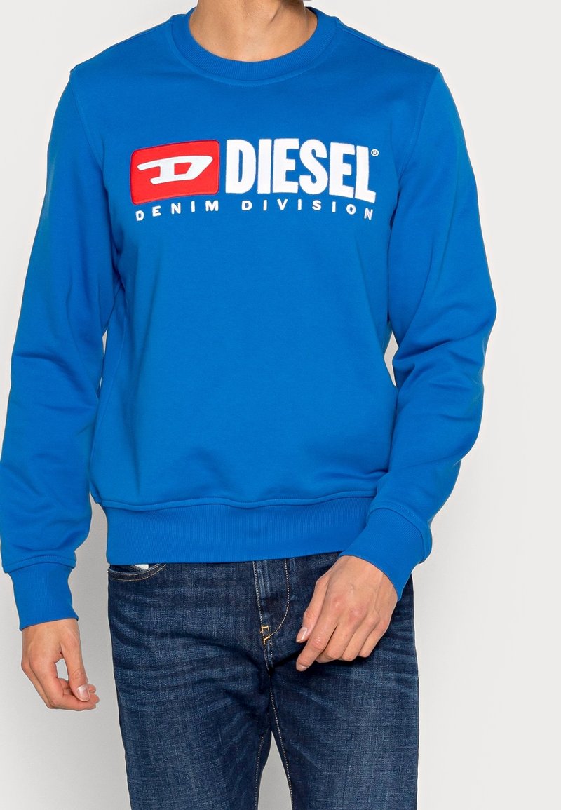 Homme portant un sweat-shirt Diesel bleu vif avec le texte "DIESEL DENIM DIVISION" en blanc et rouge, associé à un jean bleu foncé.