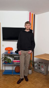 Νεαρός άνδρας με μαύρο φούτερ με κουκούλα και μπεζ παντελόνι στέκεται σε δωμάτιο με πορτοκαλί φωτιστικό, μπάλα μπάσκετ, φυτά, μεταλλικό ράφι, τηλεόραση και βαλίτσα με αυτοκόλλητα.