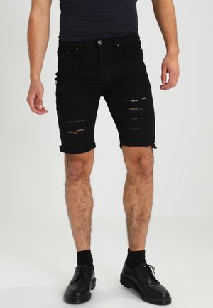 Homme portant un short en jean noir déchiré retroussé, des chaussettes noires, des chaussures habillées noires et une chemise bleu marine foncé, debout devant un fond uni.