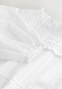 Vestido blanco texturizado con escote fruncido, mangas elásticas y un intrincado detalle de encaje a lo largo del corpiño.