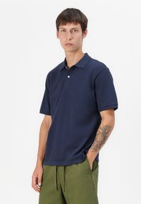 Mørkeblå polo-skjorte med korte ermer, klassisk krage og knappeplacket, sammen med olivengrønne shorts. Glatt stofftekstur.