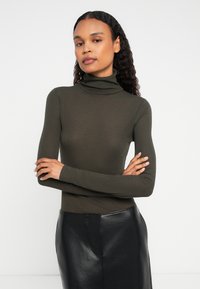 Kobieta z założonymi rękami w oliwkowym swetrze wysokim i czarnej skórzanej spódnicy, stojąca na tle jednolitego białego tła.