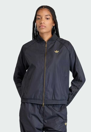 Kobieta z plecionymi włosami ubrana w czarny dres Adidas ze złotymi logo i zamkiem, ręce za plecami, neutralny wyraz twarzy.