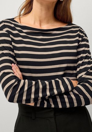 Femme aux longs cheveux portant un t-shirt à manches longues rayé horizontalement noir et beige, les bras croisés sur la poitrine.